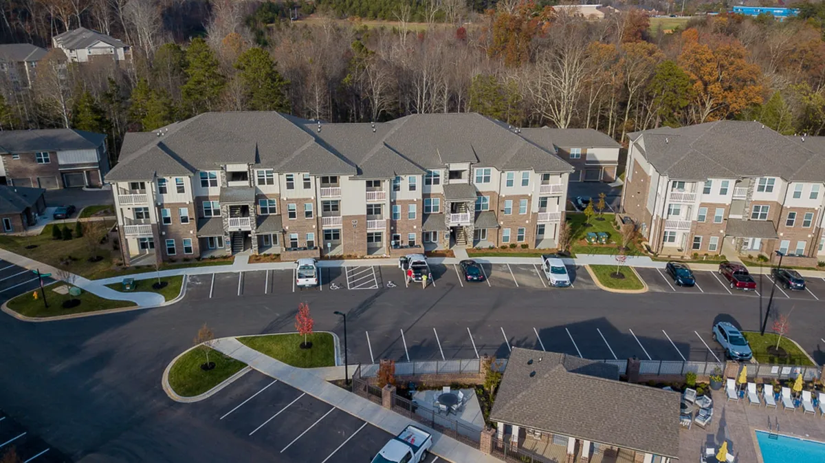 A sweeping view of the apartment complex’s exterior at sunset, featuring modern apartment buildings, a spacious parking area, a serene swimming pool, and landscaped gardens with walking trails. Residents can be seen relaxing by the pool and strolling through lush greenery.