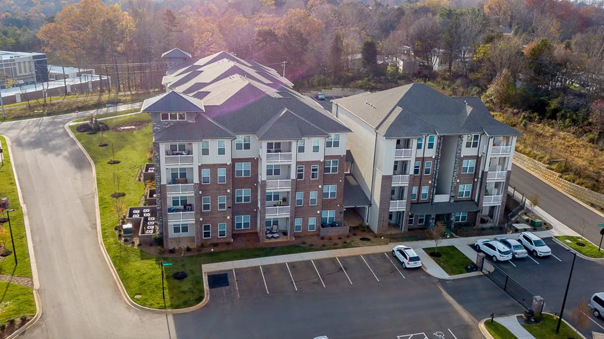 This image features a modern apartment building set amidst a well-manicured lawn bordered by mature trees and a parking area. The inviting green space and surrounding foliage speak to a peaceful, family-friendly environment, while the building’s design suggests contemporary comfort. The scene captures a sense of community, outdoor enjoyment, and a connection to nature.