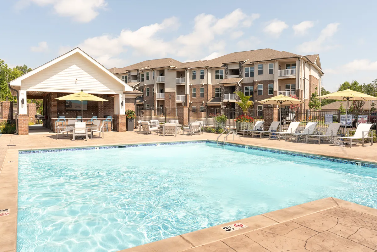The hero image showcases a sparkling swimming pool, surrounded by modern apartment buildings and lush greenery. Poolside cabanas, ample lounging chairs, and umbrella-covered tables suggest a space perfect for relaxation and socializing. The setting is bathed in warm sunset light, enhancing the inviting atmosphere.