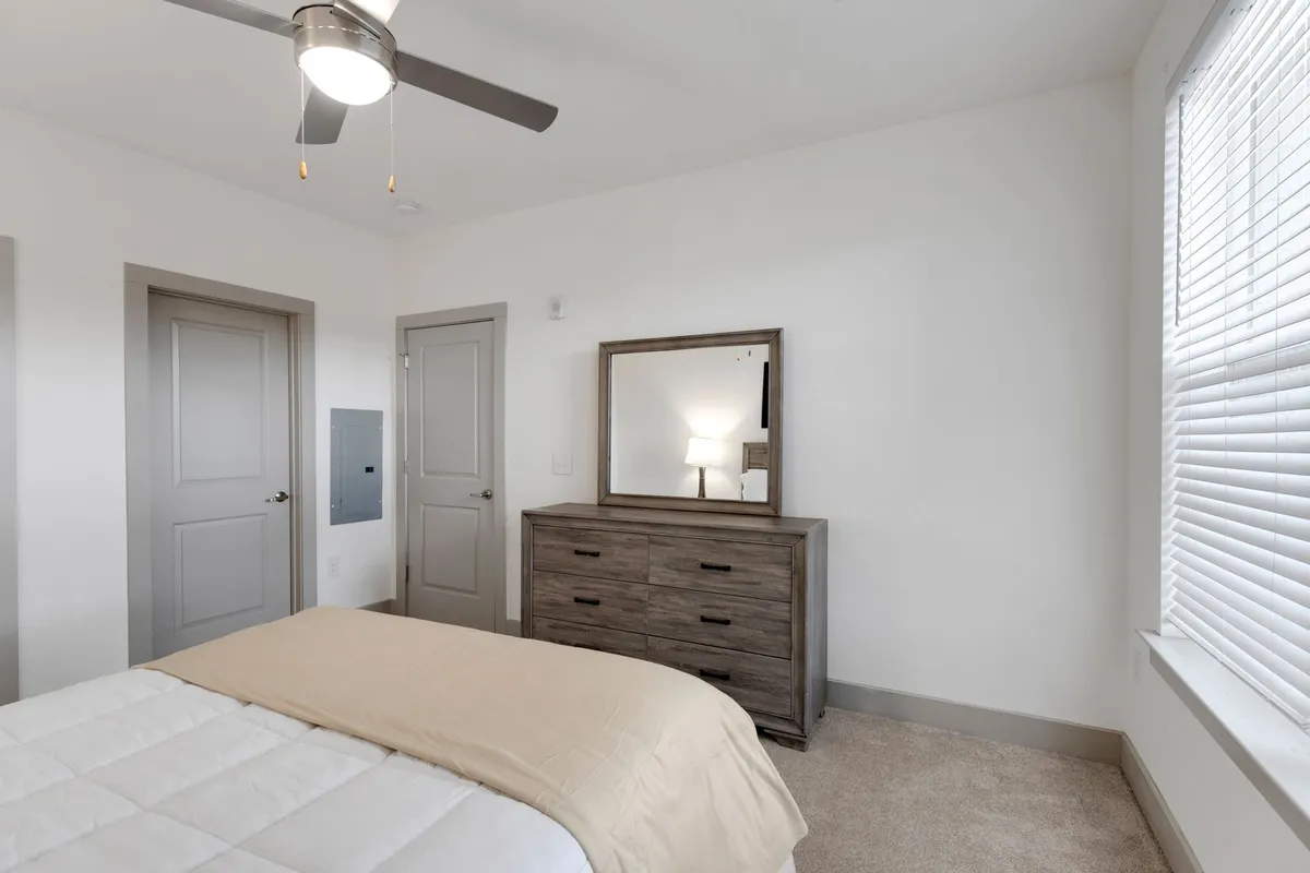 A serene, sunlit bedroom featuring a bed with cream-colored bedding, a wooden dresser, and a mirror that reflects soft, natural light. The overall arrangement and décor create an inviting and restful atmosphere.