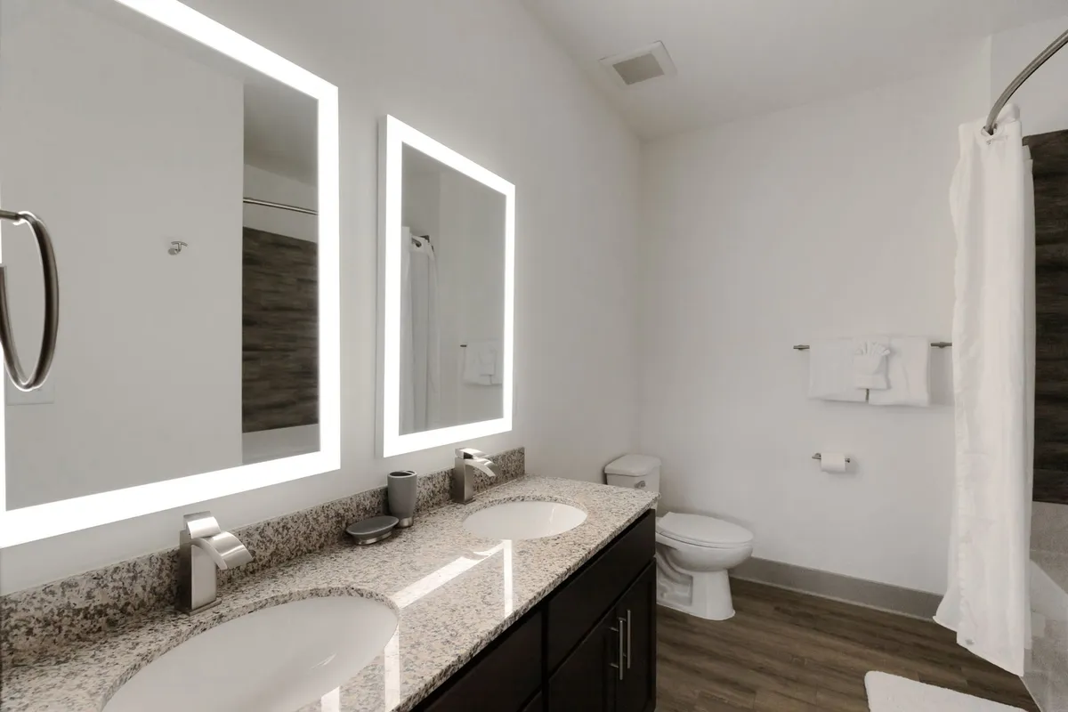 A contemporary bathroom with a double sink set in a granite countertop, backlit mirrors, and modern fixtures. The clean lines and organized layout suggest a spa-like environment ideal for morning routines or evening relaxation.