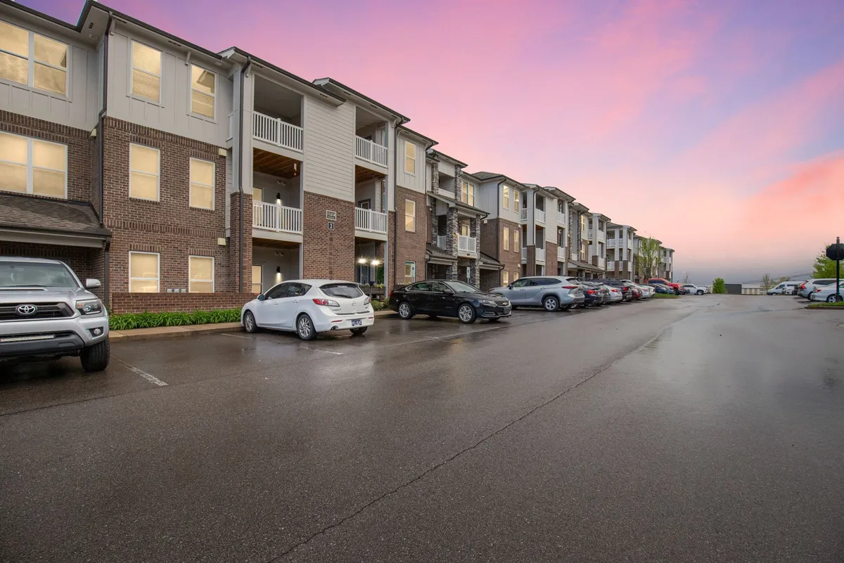 An exterior shot featuring contemporary apartment buildings, a well-maintained parking area, and lush green spaces where residents socialize and families gather. The sunset casts a warm glow, adding to the inviting atmosphere.