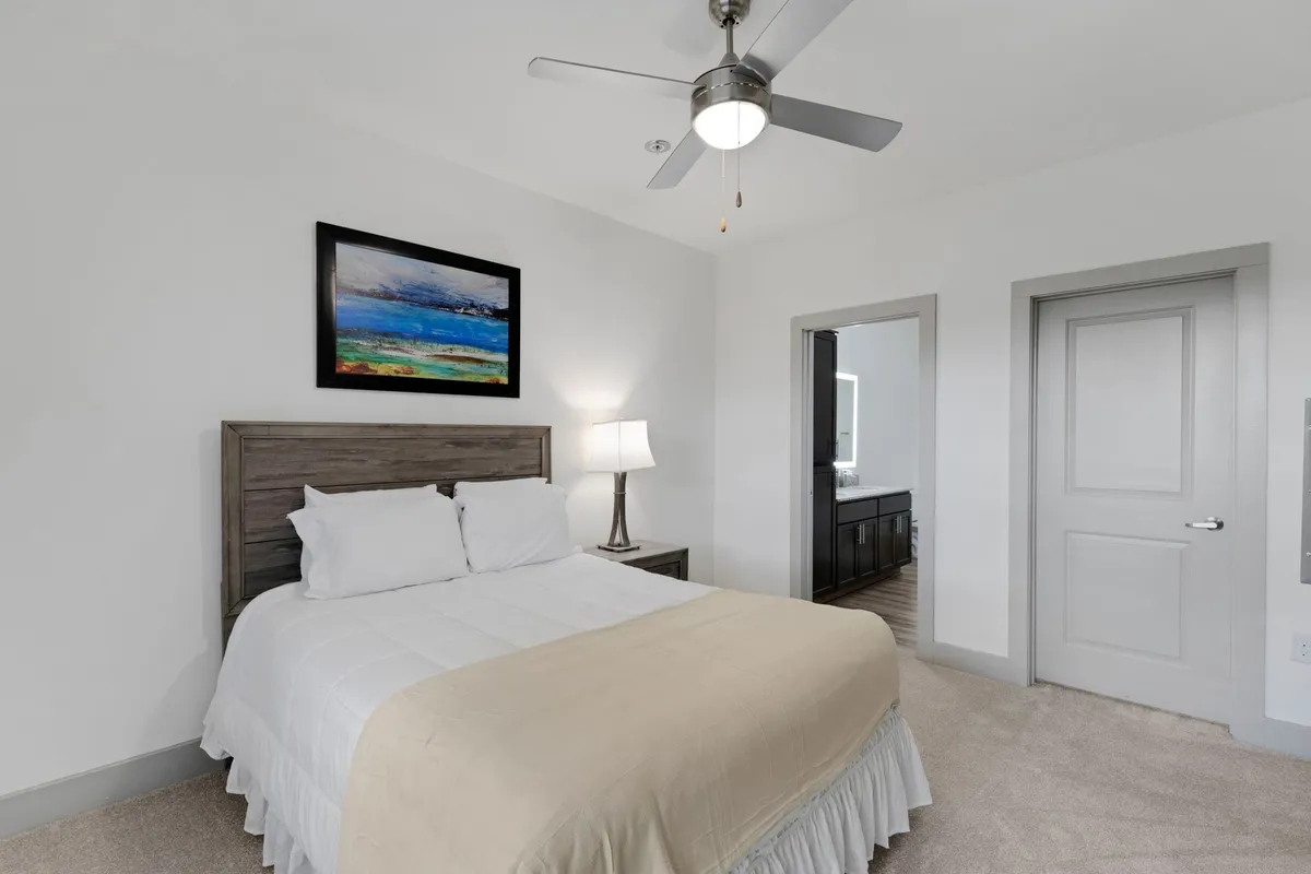 A tranquil bedroom interior showcasing a neatly made bed, soft lighting from a bedside lamp, tasteful artwork, and a partially open doorway. The room feels peaceful and private, suggesting a retreat for quiet reflection or restful breaks during a workday.