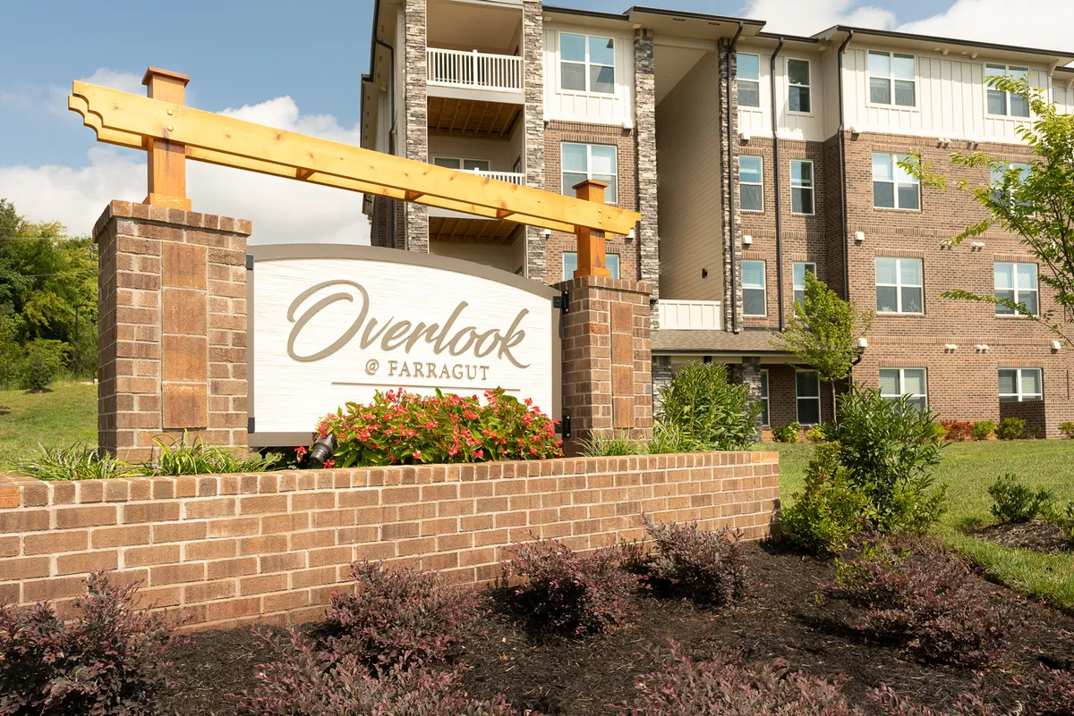 The entrance to Overlook @ Farragut is marked by a modern sign, vibrant flower beds, and the welcoming facade of the building, inviting residents and guests alike. The landscaped surroundings and clean architecture convey a professional yet homey atmosphere.