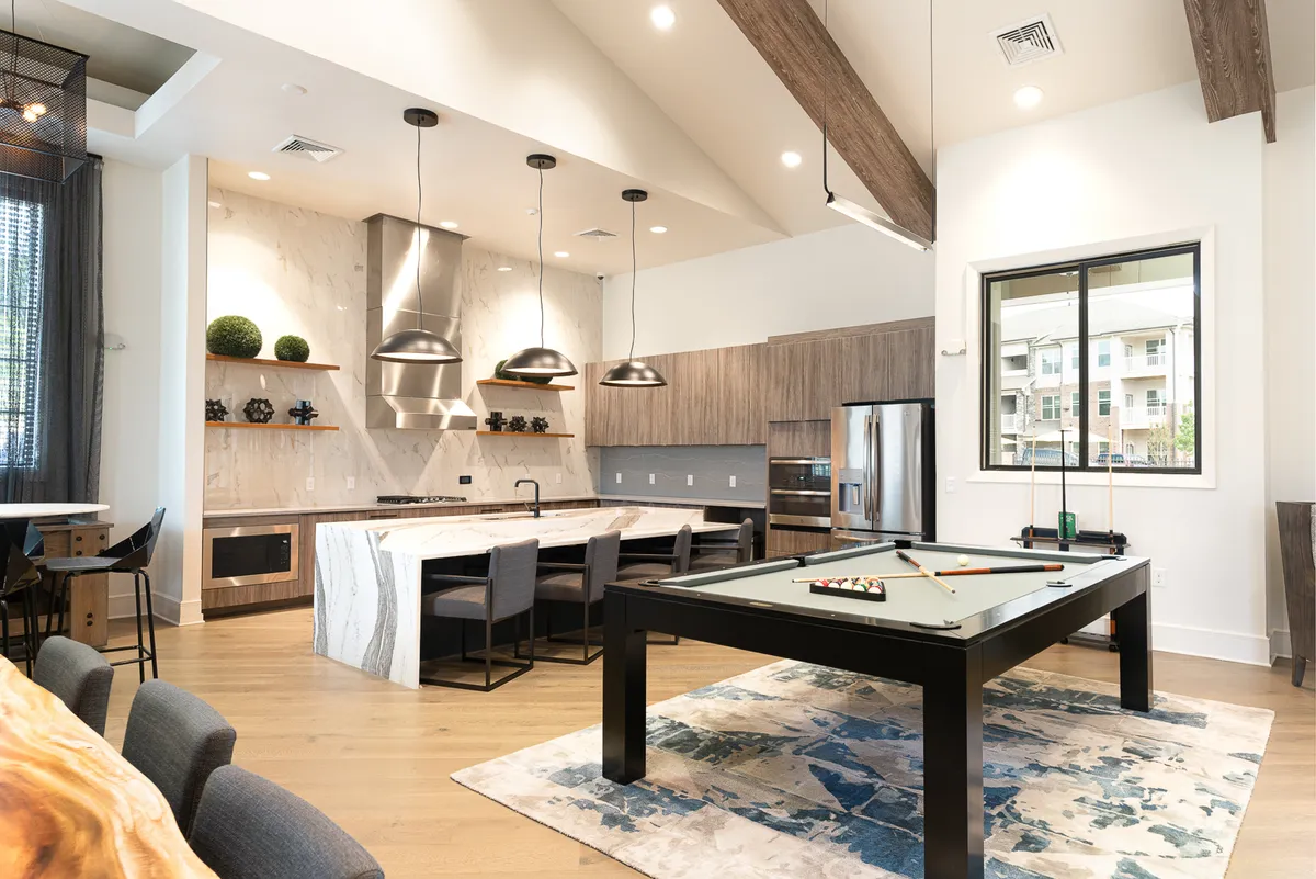 Offering a broader view, this image presents an open-plan living space with a kitchen island, dining area, and a pool table. Large windows provide plenty of natural light, and the layout encourages socializing and gathering. The kitchen and dining spaces are seamlessly integrated into the living area, supporting an interactive and hospitable environment.